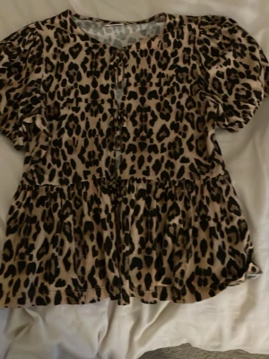 Leopardmönstrad blus med puffärm - Säljer en beige och svart leopardmönstrad blus med korta puffärmar och volang nedtill. Blusen har rund halsringning och knytning framtill. Perfekt för dig som vill sticka ut med ett djurmönster.