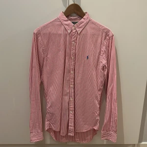 Rosavit randig skjorta från Polo Ralph Lauren. - Säljer en rosa- och vitrandig skjorta från Polo Ralph Lauren. Skjortan har lång ärm, klassisk button-down krage och blå broderad logga på bröstet. Tillverkad i bomull och har en slim fit passform. Perfekt för dig som gillar stilrena och tidlösa plagg. Skriv vid minsta lilla intresse🤩