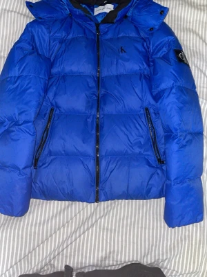 Blå pufferjacka från Calvin Klein Jeans - Säljer en blå pufferjacka från Calvin Klein Jeans med huva och svarta detaljer. Jackan har dragkedja framtill, två sidofickor med dragkedja och CK-logga på bröstet och ärmen. Perfekt för kalla dagar och riktigt snygg färg som sticker ut. Avtagbar huva. 