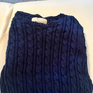 Mörkblå kabelstickad tröja från Iconic Sport - Säljer en mörkblå kabelstickad tröja från Iconic Sport i storlek medium. Tröjan har klassiskt flätmönster och rund halsringning, perfekt för kyligare dagar.