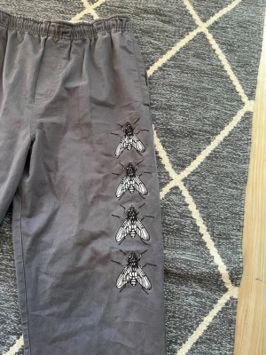Grå byxor med flugtryck och resår - Säljer ett par grå byxor från HUF med elastisk midja och raka ben. På ena benet finns fyra stora tryck av flugor i svartvitt, vilket ger en unik och edgy vibe. Byxorna har bakficka med HUF-logga och är gjorda i bomull.