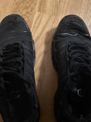Svarta Air Max Plus/Nike TN  - Säljer ett par helsvarta Air max plus/Nike TN i storlek 43 sneakers med mesh-ovandel och snygga vågformade detaljer. Skorna har snörning och en chunky sula som ger grym streetstil. Perfekta för dig som gillar en sportig look och vill ha bekväma skor till vardags.