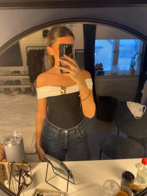 Svart och vit offshoulder body - Säljer en snygg offshoulder body i svart med vit omlott-detalj över axlarna och en guldfärgad metallspänne framtill. Toppen är tight och figurnära, perfekt att styla med jeans eller kjol. Materialet känns slätt och stretchigt. köpt på zara