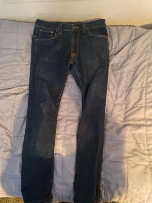 Mörkblå nudie jeans - Säljer ett par mörkblå jeans från Nudie Jeans med klassiska kontrastsömmar och snygga bakfickor med vågig brodyr. Modellen har normal passform och raka ben. Tillverkade i slitstarkt jeansmaterial av bomull. Perfekta till en avslappnad streetstil.