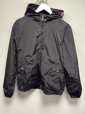 Svart vindjacka från Moncler med huva - Svart Moncler vindjacka med huva och dragkedja framtill. Jackan har två fickor med tryckknappar och elastiska muddar vid ärmslut och nederkant. Klassisk Moncler-logga på ärmen och diskreta röda och vita detaljer på huvan. Perfekt för blåsiga dagar. 100% äkta. Skriv för frågor!