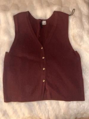 stickad väst  - Säljer en vinröd stickad väst från Divided med v-ringning och guldfärgade knappar framtill. Jag är storlek S och på mig sitter den lite löst men bekvämt. Västen har en croppad passform och är perfekt att styla över en skjorta eller t-shirt för en trendig look.