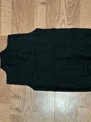 Svart stickad väst med polokrage - Säljer en svart stickad väst med hög polokrage. Västen har ett diskret ribbat mönster framtill och är ärmlös, perfekt att styla över en skjorta eller t-shirt. Enkel och clean design som funkar till många olika looks.