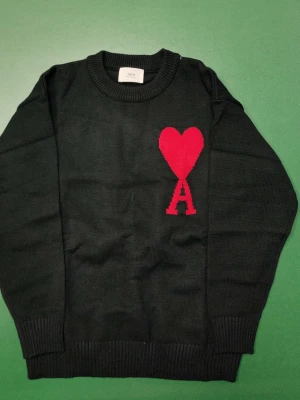 Svart stickad tröja från Ami Paris - Svart stickad tröja från Ami Paris med röd hjärtlogga och bokstaven A på bröstet. Klassisk rund halsringning och ribbade muddar vid ärmslut och nederkant. Perfekt för dig som gillar stilrena plagg med en twist.