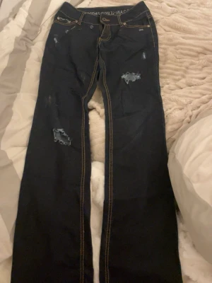 Lågmidjade Mörkblåa jeans med slitningar från Vila - Säljer ett par mörkblåa jeans från Vila Clothes med coola slitningar och råa detaljer på benen. Jeansen har kontrastsömmar och klassisk femficksdesign. Passar dig som gillar en edgy stil och vill ha något som sticker ut i garderoben.