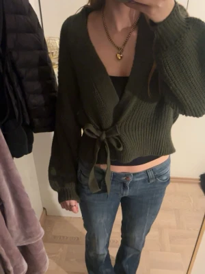 Mörkgrön stickad omlottkofta - Säljer en mörkgrön stickad omlottkofta med knytband i midjan. Koftan har breda, långa ärmar och ribbade muddar. Croppad modell som ger en snygg siluett och passar perfekt till jeans eller kjol.