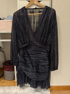 Glittrig svart kortklänning från Maje - Supersnygg svart kortklänning från Maje med glittriga ränder och v-ringning. Klänningen har långärmade ärmar, omlottkänsla och volangdetalj nedtill. Materialet är skimrande och ger en lyxig känsla, perfekt för att sticka ut.