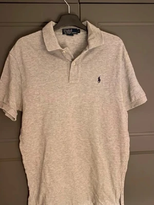 Grå pikétröja från Polo Ralph Lauren - Klassisk grå pikétröja från Polo Ralph Lauren med korta ärmar och liten broderad logga på bröstet. Tröjan har krage och knappar framtill. Perfekt basic till garderoben och enkel att matcha.