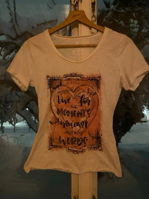 Vit t-shirt med tryck från Gina Benotti - Vit t-shirt från Gina Benotti med rund hals och korta ärmar. Framsidan har ett stort tryck med texten 'Live for the moments you can't put into words' i ett hjärta, dekorerat med fjärilar och orange bakgrund. Tillverkad i mjuk bomull, perfekt för en chill look.