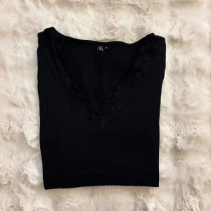 Svart långärmad topp med spets - Säljer en svart långärmad topp med djup v-ringning och snygg spetskant runt halsen. Toppen är i mjukt material och har en enkel, stilren look med fina spetsdetaljer som ger en extra touch.
