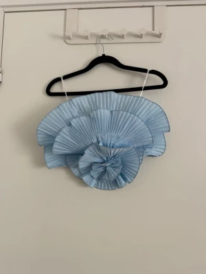 Ljusblå plisserad bandeau topp - Unik ljusblå bandeau topp med tunna axelband och stora plisserade volanger framtill som ger en cool och extravagant look. Toppen har en strukturerad form och är perfekt för dig som vill sticka ut. Materialet känns lätt och har en snygg glans. Använd en gång.
