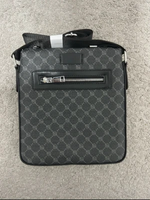 Svart axelväska med monogrammönster - Snygg svart axelväska med klassiskt monogrammönster och silverfärgad dragkedja framtill. Väskan har justerbar axelrem och är tillverkad i skinn och syntet. Perfekt för dig som vill ha en stilren och praktisk väska med exklusiv känsla. Dens dragkedja där framme är lite sönder och väldigt svår att öppna och stänga men den är helt ny. 