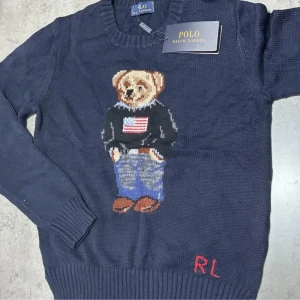 Mörkblå stickad tröja Polo Ralph Lauren - Mörkblå stickad tröja från Polo Ralph Lauren med ikonisk björn på bröstet och RL-broderi nertill. Tröjan har rund halsringning och långa ärmar. Perfekt för dig som vill ha en klassisk och snygg look med en twist.