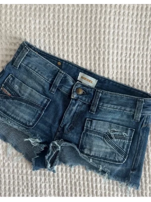 Blå jeansshorts från Diesel - Säljer ett par blå jeansshorts från Diesel med råa, fransiga kanter och slitna detaljer. Klassisk femficksmodell med låg midja och cool tvätt. Perfekta för sommaren och riktigt snygga till sneakers eller boots.