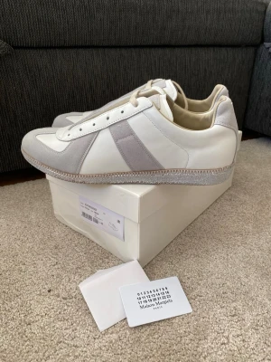 Maison Margiela GATs sneakers vit/grå - Maison Margiela sneakers i vitt och ljusgrått med klassisk retrodesign. Ovandel i skinn och mocka, beige snörning och gummisula med logotyp under. Snygg minimalistisk look och diskreta detaljer, perfekt för dig som gillar stilrena sneakers.