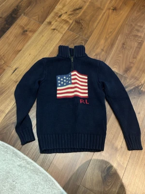 Mörkblå stickad tröja Polo Ralph Lauren - Mörkblå stickad tröja från Polo Ralph Lauren med amerikansk flagga framtill och RL-broderi. Tröjan har hög krage, half zip-dragkedja och ribbade muddar. Perfekt för dig som vill ha en klassisk och snygg look med lite preppy känsla.