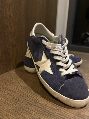 Blå Golden Goose skor - Snygga blå sneakers från Golden Goose med vit stjärndetalj på sidan och vita snören. Ovandelen är i mocka och sulan är vit med svart undersida. Skorna har en klassisk låg profil och cool streetkänsla. Är villig att gå ner lite i pris.