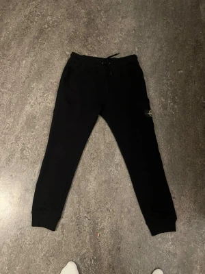 Svarta mjukisbyxor från Stone Island - Säljer ett par svarta mjukisbyxor från Stone Island med klassisk patch på benet. Byxorna har dragsko i midjan, ribbade muddar vid bensluten och en praktisk ficka på sidan. Tillverkade i mjukt bomullsmaterial för maximal komfort.