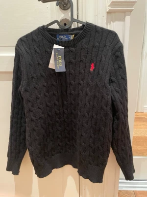 Svart kabelstickad tröja Polo Ralph Lauren - Kabelstickad svart tröja från Polo Ralph Lauren med klassisk röd logga broderad på bröstet. Rund halsringning och ribbade muddar vid ärmslut och nederkant. Perfekt för dig som gillar stilrena och tidlösa plagg.