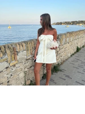 Klänning zara - Superfin axelbandslös vit miniklänning med stor rosettdetalj framtill och långa band som hänger ner. Klänningen har en stilren och elegant look med rak passform och är perfekt för sommarens alla tillfällen.