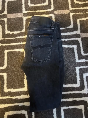 Svarta slim jeans från Nudie Jeans - Säljer ett par svarta slim jeans från Nudie Jeans med klassisk femficksdesign och snygg Nudie logga på bakfickan. Jeansen har en smal passform och är tillverkade i mjukt jeansmaterial som sitter snyggt längs benen. Perfekta för dig som gillar en clean och stilren look. Storlek 27 32