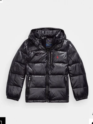 Svart pufferjacka från Polo Ralph Lauren - Säljer en svart pufferjacka från Polo Ralph Lauren med huva och blank finish. Jackan har dragkedja framtill, två sidofickor med dragkedja och den klassiska röda loggan på bröstet. Perfekt för kalla dagar och riktigt snygg till alla outfits. Storlek XL BARN vilket ungefär motsvarar storlek S, köpt på kidsbrandstore. I perfekt skick, använd ett fåtal gånger