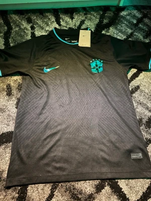 Svart Brasil Nike fotbollströja - Säljer en svart Nike fotbollströja för Brasilien med turkosa detaljer och emblem på bröstet. Tröjan har korta ärmar, rund hals med turkos kant och är tillverkad i lätt polyester med diskret mönster i tyget. Perfekt för dig som älskar fotboll och vill sticka ut på planen.