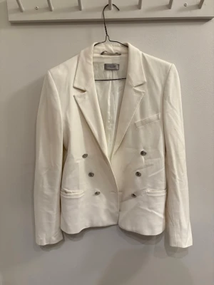 Vit kavaj med silvriga knappar - Snygg vit kavaj från Hunkydory med dubbelknäppning och silvriga knappar framtill. Klassisk krage, långärmad och med fickdetaljer. Perfekt för dig som vill ha en stilren och fräsch look.