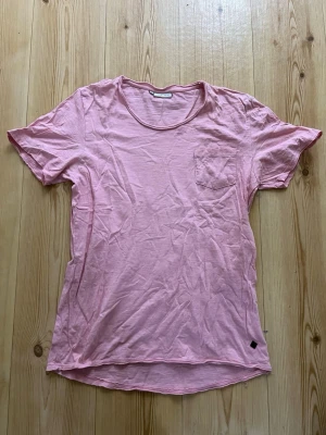 Rosa t-shirt från Casual Friday - En enkel rosa t-shirt från Casual Friday i storlek L. T-shirten har rund hals, korta ärmar och en liten bröstficka. Tillverkad i mjuk bomull som känns skön mot huden. Perfekt för en chill och avslappnad stil.