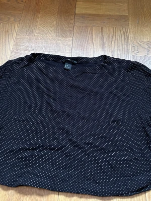 Svart prickig blus från H&M - Säljer en svart blus från H&M i storlek 38 med små vita prickar över hela tyget. Blusen har en klassisk och enkel form, perfekt att styla med jeans eller kjol. Materialet känns lätt och luftigt.