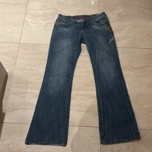 BH Jeans blå broderade bootcut jeans - Säljer ett par blå jeans från BH Jeans med snygga broderade detaljer i turkos, orange och vitt på fickorna. Jeansen har bootcut passform, nitar vid linningen och klassisk femficksdesign. Materialet är jeans med slitningar för en cool look. PRISET KAN DISKUTERAS!!