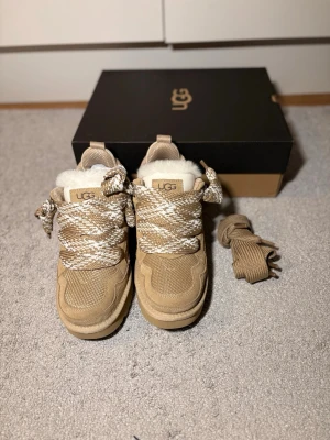 Uggs lowmel  - Snygga beige sneakers från UGG med breda, mönstrade snören i beige och vitt. Skorna har en robust sula, detaljer i mocka och mesh samt fluffigt vitt fårfoder på insidan för extra värme. Perfekta för kyligare dagar och riktigt sköna att bära.