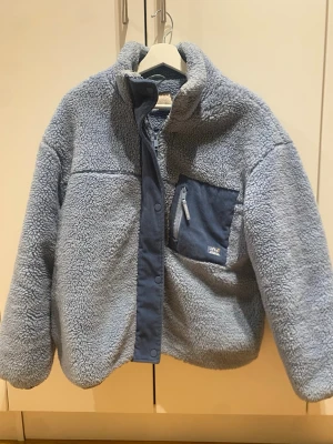 Blå teddyjacka från pull and bear  - Mysig blå teddyjacka från pull and bear  med hög krage och en praktisk bröstficka med dragkedja. Jackan har en stängning med både dragkedja och tryckknappar framtill. Perfekt för kyliga dagar och har en riktigt skön oversized känsla. Priset går att diskutera! 