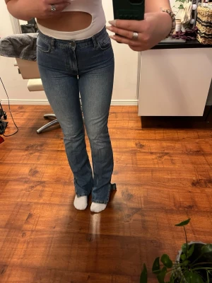 Jeans med slits - Säljer ett par klassiska jeans i blå tvätt med bootcut-modell och slit nertill. Jeansen är mid waist och har raka ben som blir lite utsvängda nertill. Tillverkade i slitstarkt jeanstyg med fem fickor