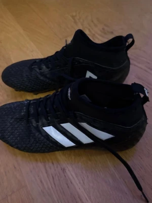 Svarta Adidas fotbollsskor med snörning - Säljer ett par svarta Adidas fotbollsskor med vita ränder på sidorna och högre skaft. Skorna har snörning och en texturerad yta för bättre bollkontroll. Perfekta för dig som vill ha stilrena och funktionella skor på planen.