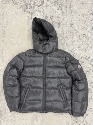 Grå moncler maya - Säljer en svart pufferjacka från Moncler med stor huva och dragkedja framtill. Jackan har två snedställda fickor med dragkedja och klassisk Moncler-logga på ärmen. Insidan har ett färgglatt serietryck. Perfekt för kalla dagar och riktigt snygg streetstil.