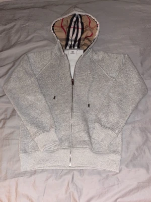 Grå Burberry Zip Up hoodie helt ny  - Grå zip hoodie från Burberry i storlek XS. Klassisk och stilren modell med dragkedja framtill, justerbar huva med Burberrys ikoniska rutmönster på insidan samt diskret broderad logotyp på bröstet. Mjuk och bekväm kvalitet, perfekt för vardagsbruk. Och sprillansny , 