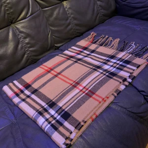 Burberry halsduk - Säljer en Burberry halsduk med klassiskt rutigt mönster i svart, vitt och rött. Halsduken har snygga fransar i ändarna och är mjuk och skön att ha runt halsen. Perfekt accessoar för att lyfta din outfit.