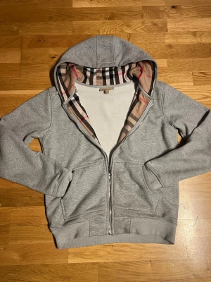 Grå zip-hoodie från Burberry - Snygg grå hoodie med dragkedja från Burberry. Klassisk huva med det ikoniska rutmönstret på insidan, två fickor fram och ribbade muddar. Perfekt för en avslappnad och stilren look.