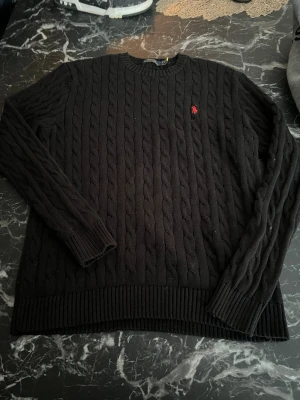 Svart kabelstickad tröja Polo Ralph Lauren - Svart kabelstickad tröja från Polo Ralph Lauren med klassisk röd broderad logga på bröstet. Tröjan har rund halsringning, långa ärmar och ribbstickade muddar. Perfekt för en stilren och tidlös look.