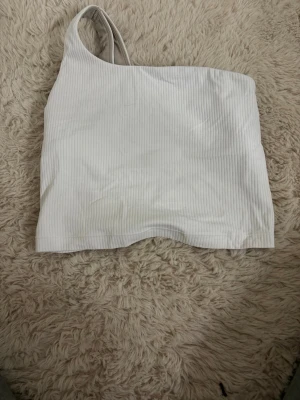 Vit ribbad oneshoulder topp Lululemon - Säljer en vit ribbad oneshoulder topp från Lululemon. Säljer på grund av att den ör för liten. Inga defekter. Diskret Lululemon-logga på sidan. Storlek 2