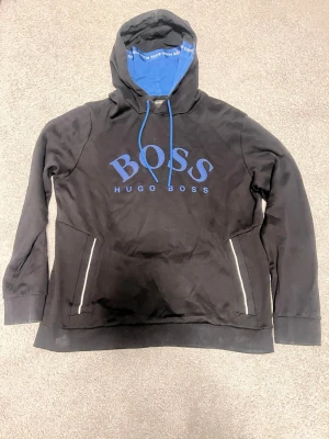 Svart hoodie från Hugo Boss - Snygg svart hoodie från Hugo Boss med stor blå logga på bröstet och blå dragsko i huvan. Insidan av huvan är blå med vit Hugo Boss-text. Hoodien har två fickor med dragkedja och är tillverkad i mjukt material för en bekväm känsla.