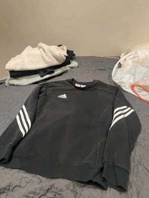 Svart Adidas sweatshirt med vita ränder - Klassisk svart sweatshirt från Adidas med vita ränder på ärmarna och Adidas-logga på bröstet. Tröjan har rund halsringning och är tillverkad i mjukt material som passar perfekt till en avslappnad stil.