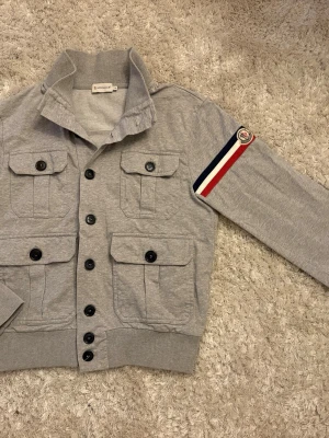 Grå overshirt från Moncler - Snygg grå overshirt från Moncler med fyra stora fickor framtill och svarta knappar. Jackan har ribbad krage och muddar samt en Moncler-logga med röd, vit och marinblå detalj på ärmen. Perfekt för dig som gillar streetwear och stilrena plagg.