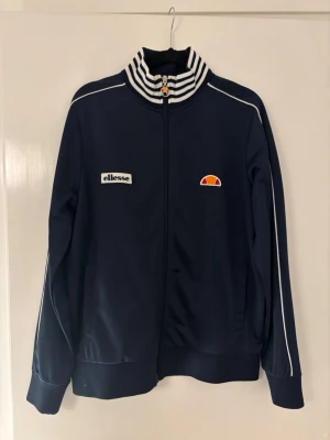 Marinblå ziptröja från Ellesse - Snygg marinblå ziptröja från Ellesse med vita detaljer och logga på bröstet. Tröjan har hög krage med svartvita ränder, vita piping längs ärmarna och muddar vid ärmslut och nederkant. Perfekt för en sportig och retro look.