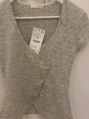 Grå omlottopp med knappar från Zara - Helt utsåld på hemsidan, Säljer en ljusgrå omlottopp från Zara med korta ärmar och stora dekorativa knappar framtill. Toppen har v-ringning och är tillverkad i mjukt stickat material som ger en skön och trendig look. Perfekt att styla med jeans eller kjol. Aldrig använd, storlek M. 